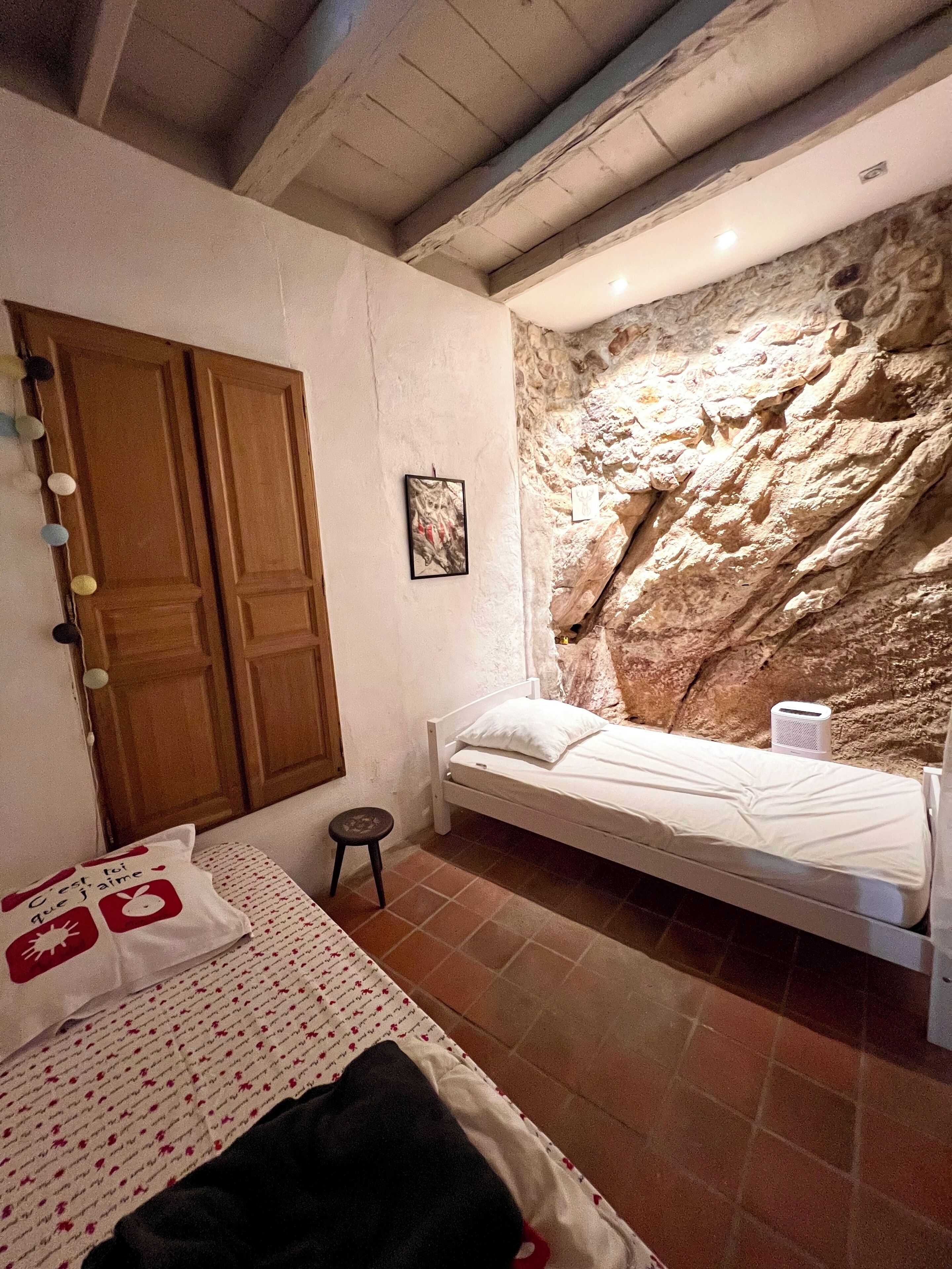 A bedroom with a bed, a small stool, a wooden door, and a wall with a rock-like texture.