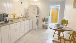 A modern kitchen with white cabinets, a built-in washing machine, a dining area with a wooden table and chairs, and a small shelf with cleaning supplies.
