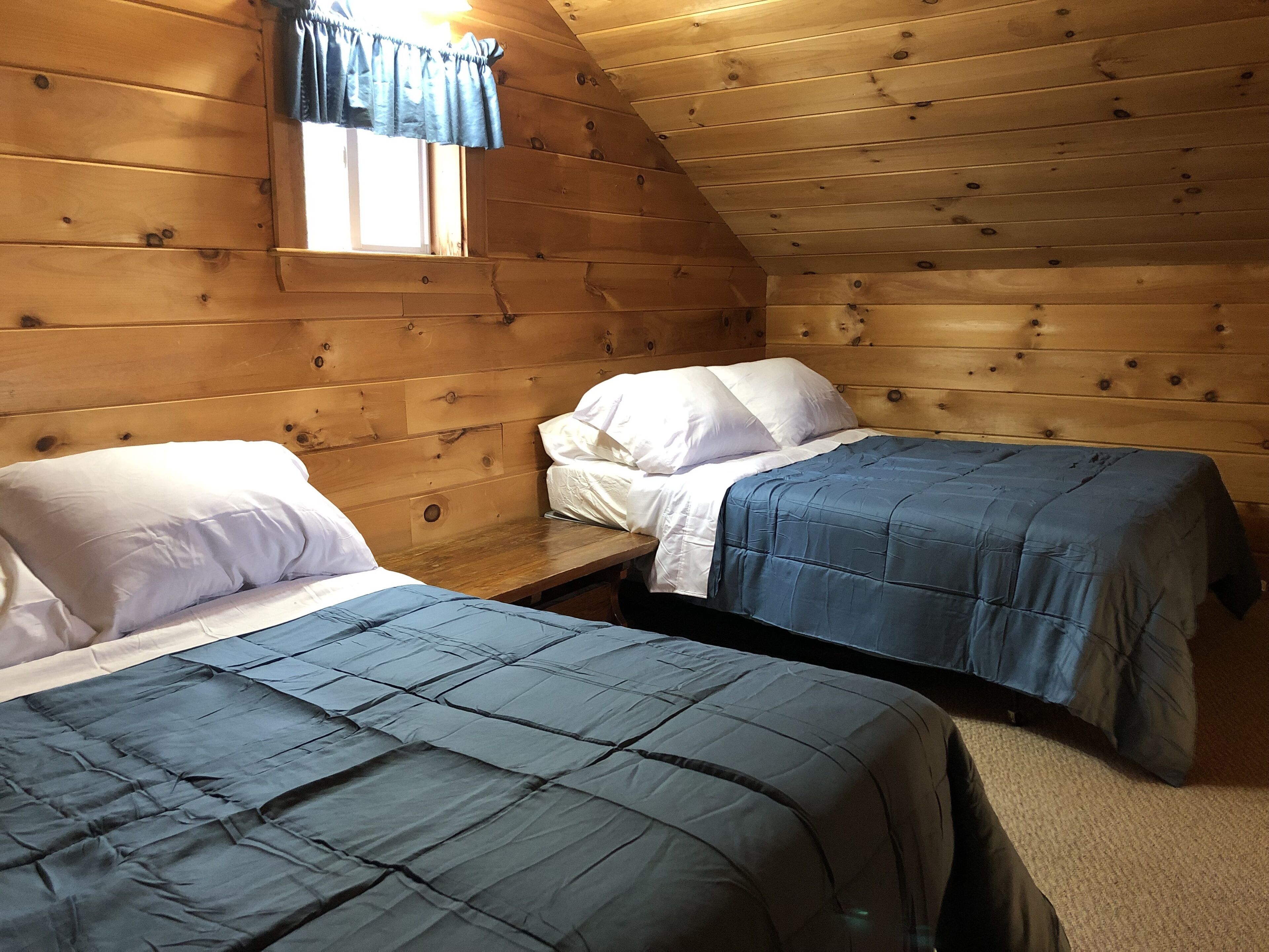A cabin with two wooden beds, a window with curtains, and a sloped ceiling.