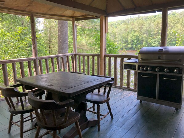 A covered outdoor area with a wooden table and chairs, a grill, and a view of a forested area. - Kennebec Riverside Cabins (West Forks)