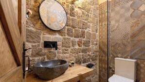 Une salle de bain avec un mur en pierre, un meuble-lavabo en bois, un grand lavabo, des toilettes et un pare-feu en carreaux décoratifs.