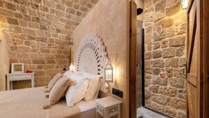 Une chambre avec un mur en pierre, un lit avec une literie blanche, une table de chevet et un luminaire fixé au mur.