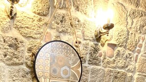 Une salle de bain aux murs en pierre, avec un miroir rond, un lavabo en pierre et une étagère en bois où sont posées des serviettes.