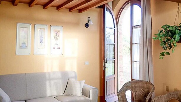 A cozy room with a wooden ceiling, a grey sofa, a wicker chair, and a large window with curtains. - Aria Apartment: Romantic Stay with Breakfast (loc Resta Buonconvento)