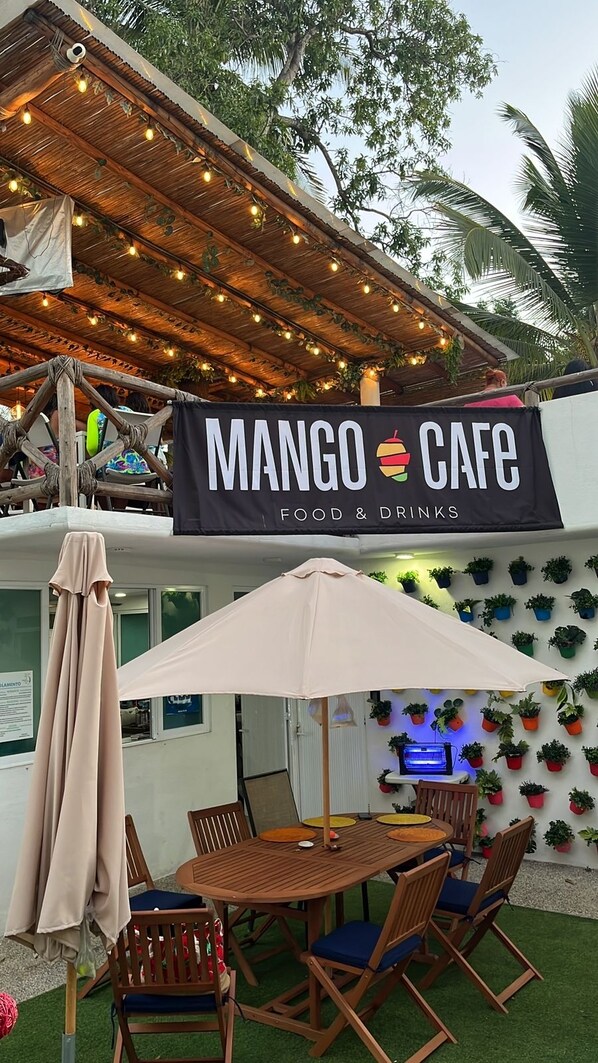 An outdoor seating area with a table and chairs, a hanging sign for Mango Cafe, and a wall decorated with potted plants.