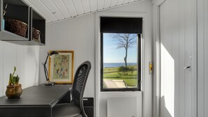 A room with a desk, chair, and a large window offering a view of the sea and a tree.