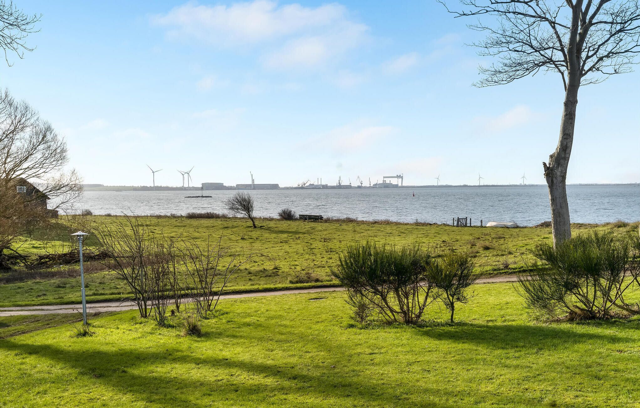 Un espace verdoyant avec un sentier, un arbre et une vue sur une étendue d’eau, où l’on aperçoit au loin des installations industrielles et des éoliennes.