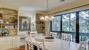 Une cuisine moderne avec des meubles blancs, une table à manger avec des chaises, un luminaire suspendu et une vue sur les arbres à travers de grandes fenêtres.