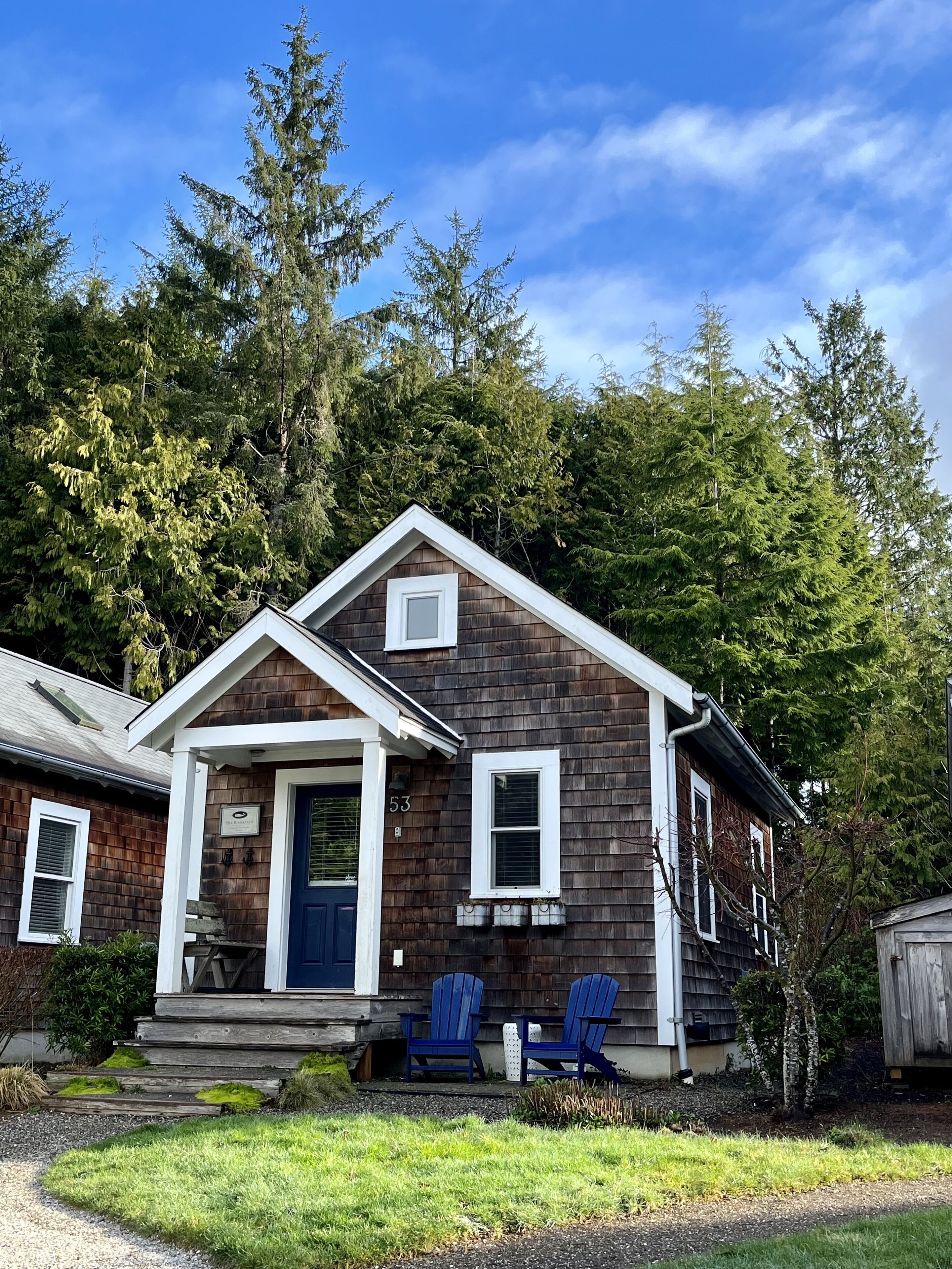 A wooden house with a blue door, numbered 53, and two blue Adirondack chairs outside.