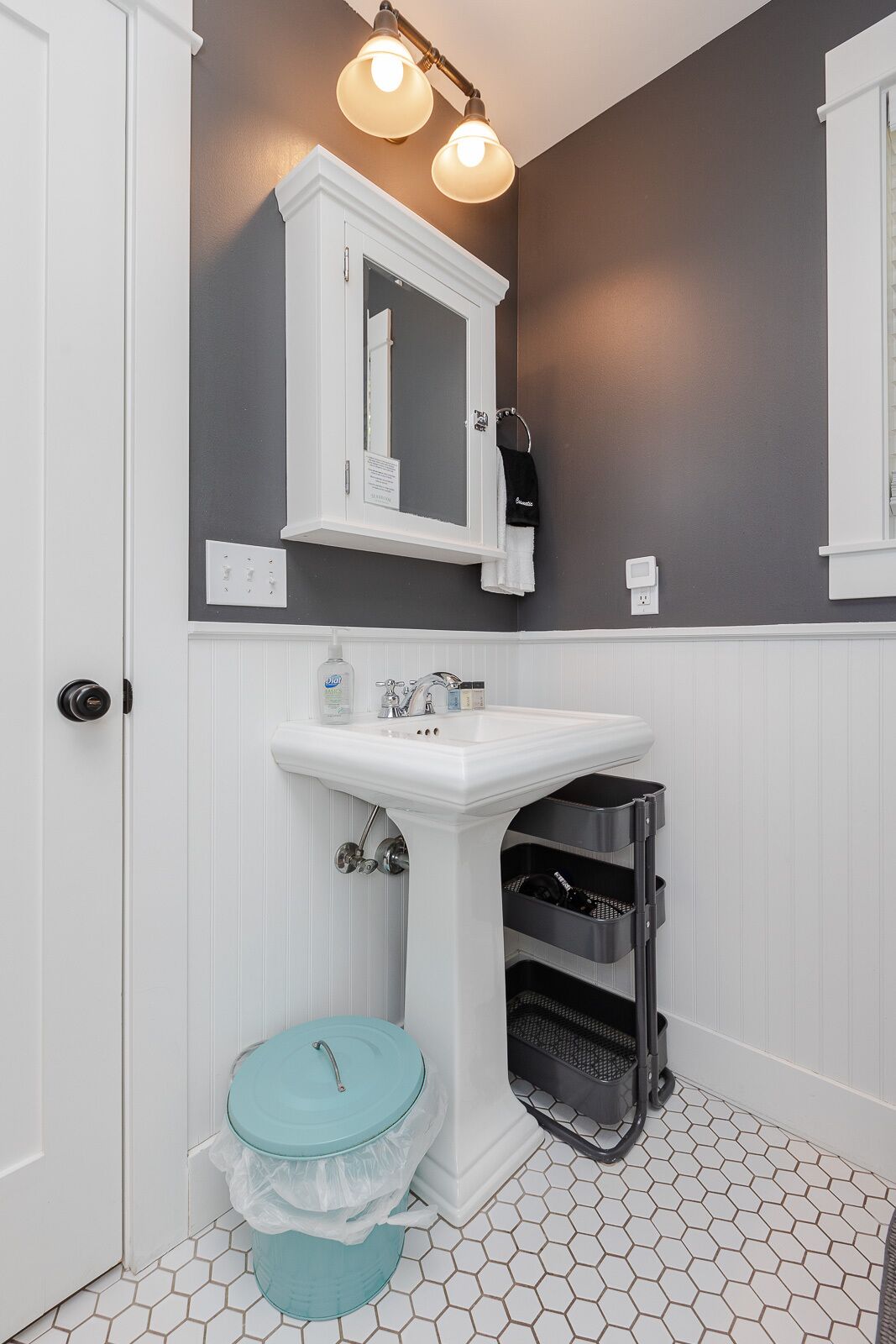 Ein Badezimmer mit einem weißen Waschbecken, einem Spiegel darüber und einer Toilette mit einem türkisblauen Deckel. Zwei Leuchten sind über dem Spiegel angebracht.
