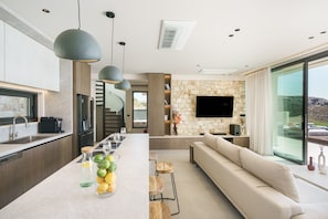 A modern kitchen with a central island, a dining area with a sofa and coffee table, and a living room with a stone wall and large TV. - Villa 7 Seas - With Amazing View (Lefkogia)