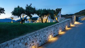 A stone wall with a paved path, trees, and a building with illuminated windows. - Villa 7 Seas - With Amazing View (Lefkogia)