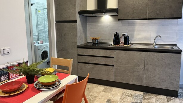 A modern kitchen with grey cabinets, a black countertop, and a stainless steel sink. There is a dining table with a red tablecloth and a potted plant. - Bnbook Studio Beatrice Rho Fiera (Rho)