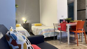 A compact room with two beds, a dining table, and a chair. There is a small table with a plant and a lamp. - Bnbook Studio Beatrice Rho Fiera (Rho)
