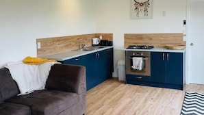 A compact living space with a kitchenette, a sofa, a dining table, and a hanging light fixture. - Country Studio Out West (Helensville)