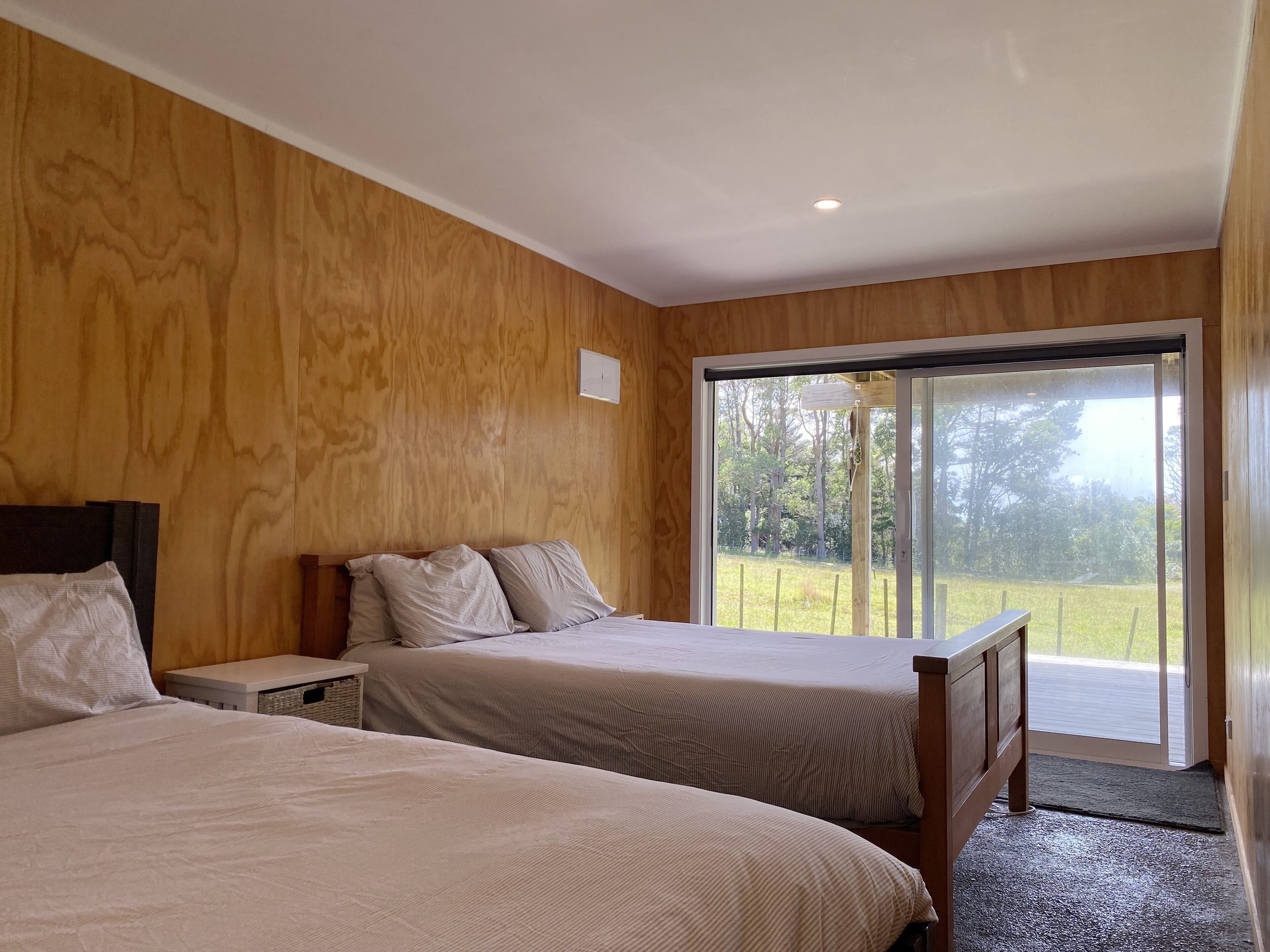 A bedroom with two beds, wooden walls, a small table, and a large sliding glass door leading to a grassy area.