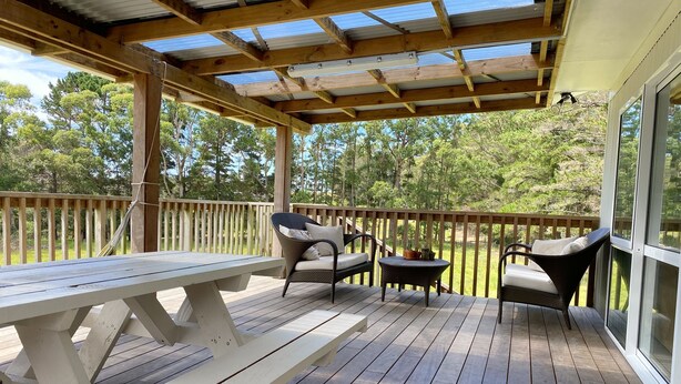 A wooden deck with a picnic table and two wicker chairs, a small round table, and a glass door leading to a garden.