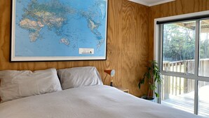 A bedroom with a large world map on the wall, a bed with striped bedding, a nightstand with a lamp, and a window with a view of a deck and trees.