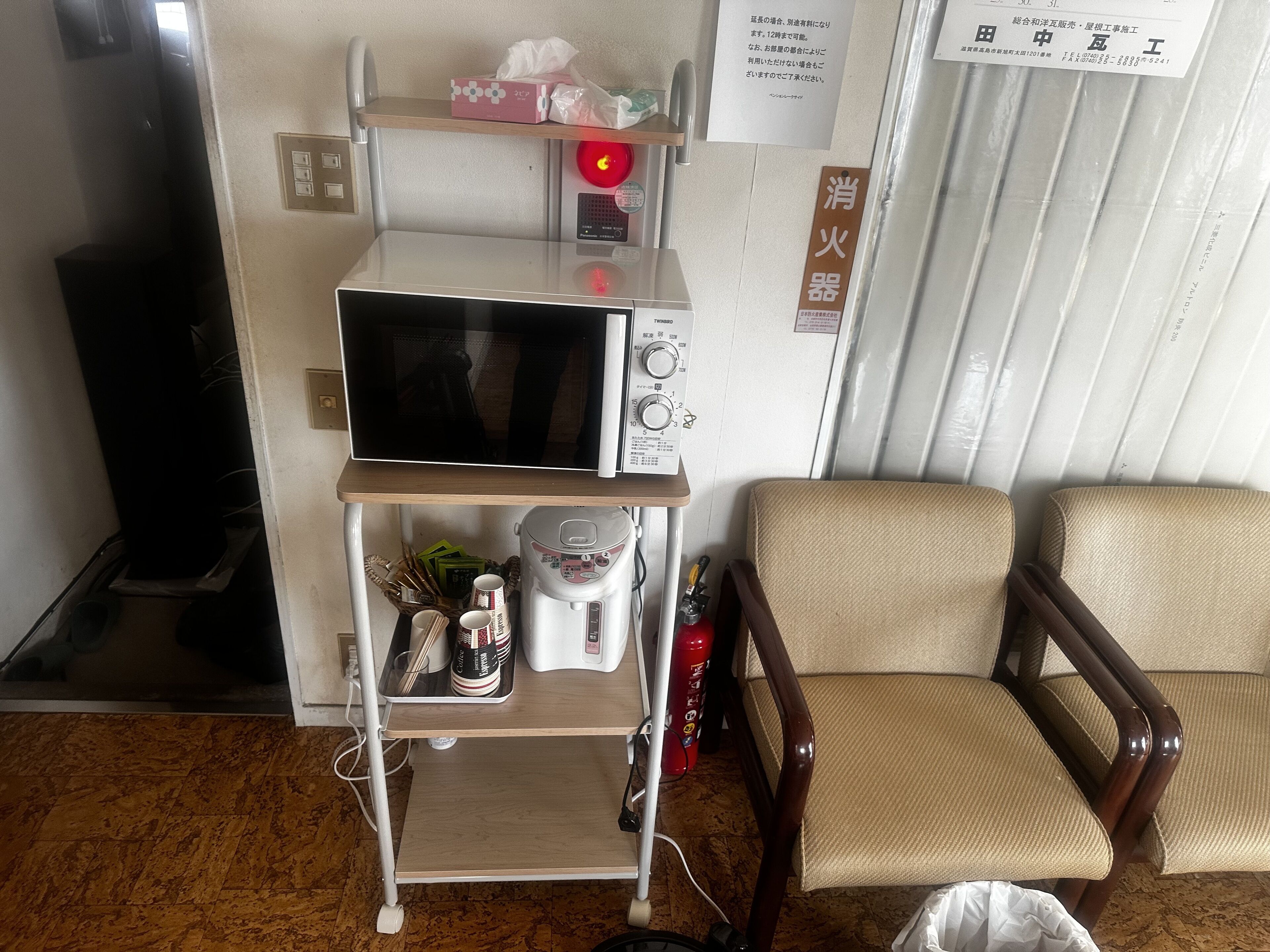 Eine Küchenzeile mit Mikrowelle, Kaffeemaschine und einer Ablagefläche mit Gegenständen. Im Zimmer befindet sich ein Stuhl und ein Feuerlöscher.
