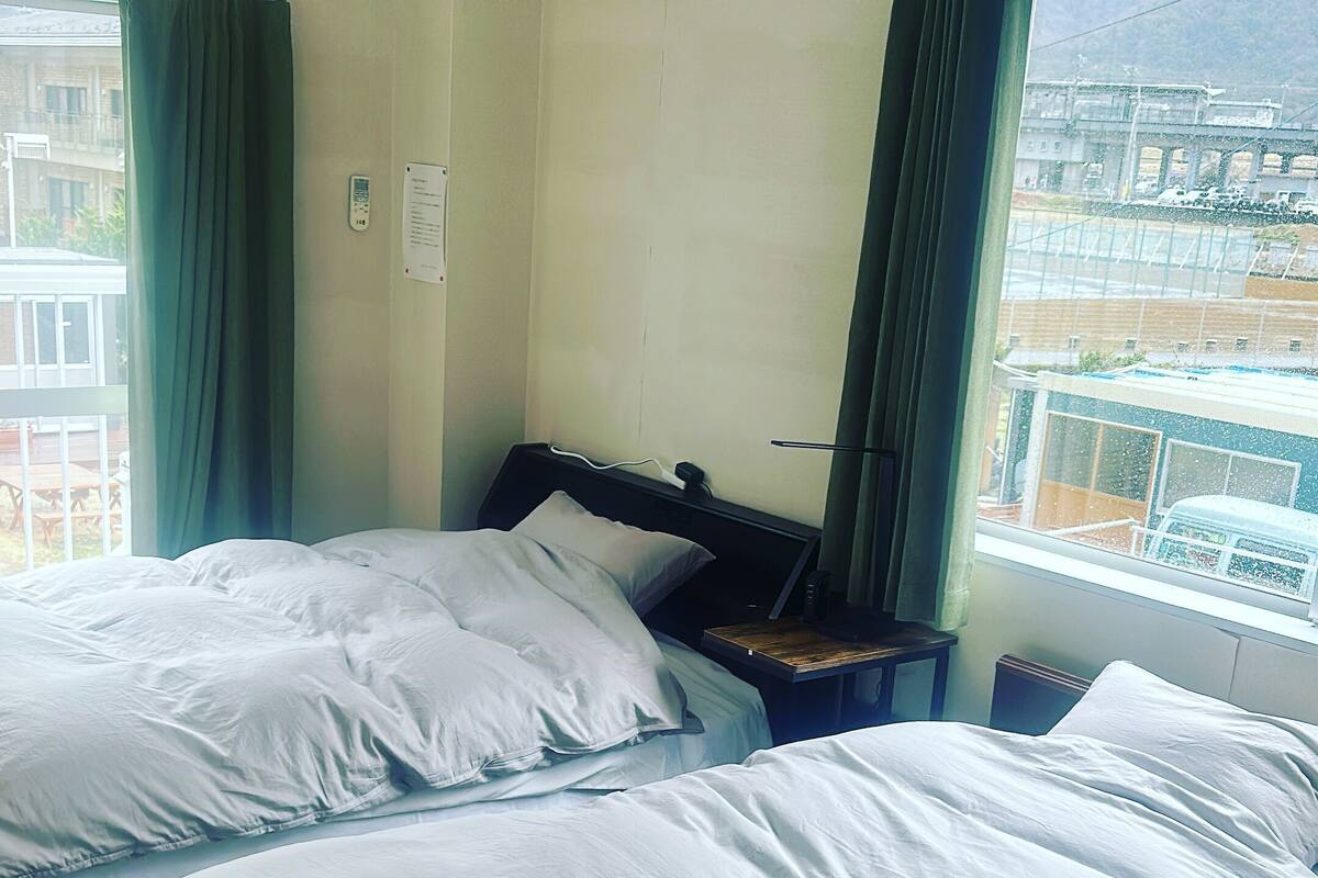 A hotel room with two beds, a bedside table, a lamp, and a window with a view of buildings.