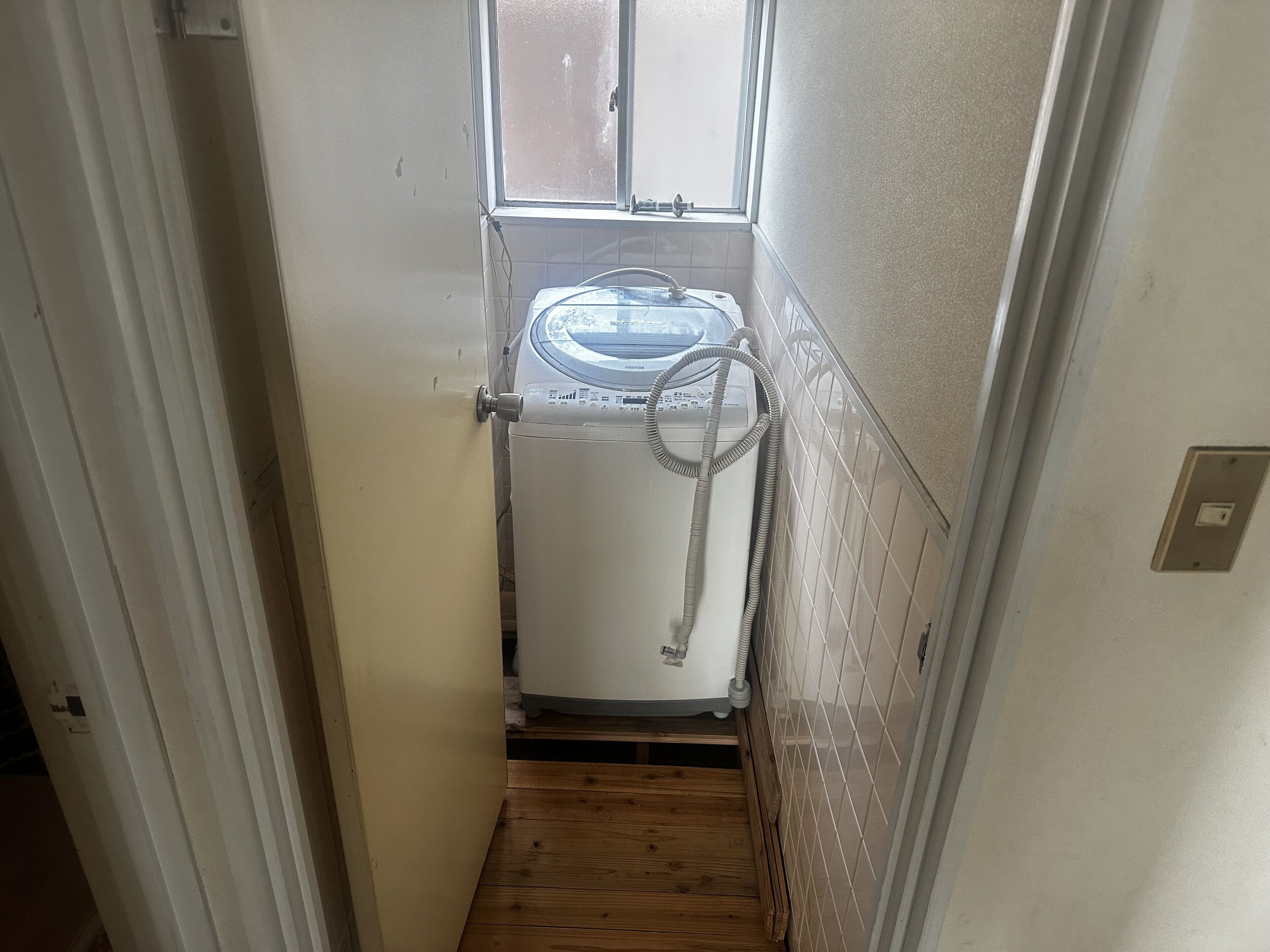 A narrow hallway with a washing machine in the corner, a window above it, and a wooden floor.