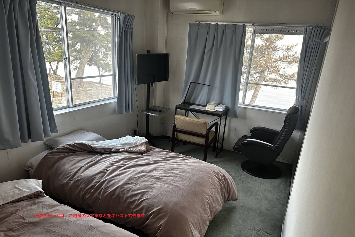 A hotel room with two beds, a desk with a computer, a chair, and large windows with curtains.