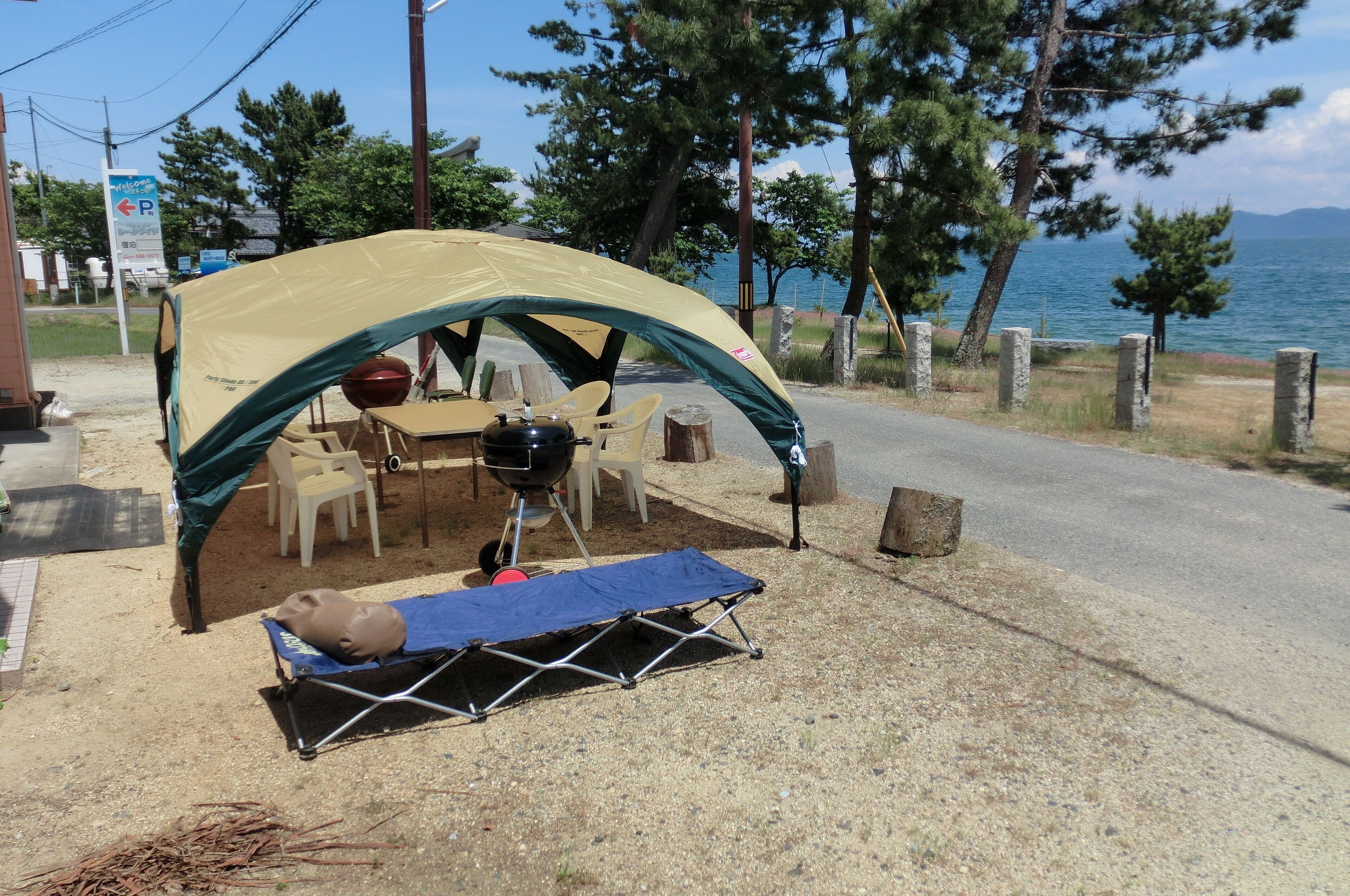 A camping setup with a tent, a table, chairs, a grill, and a sleeping mat by a road with a lake view.