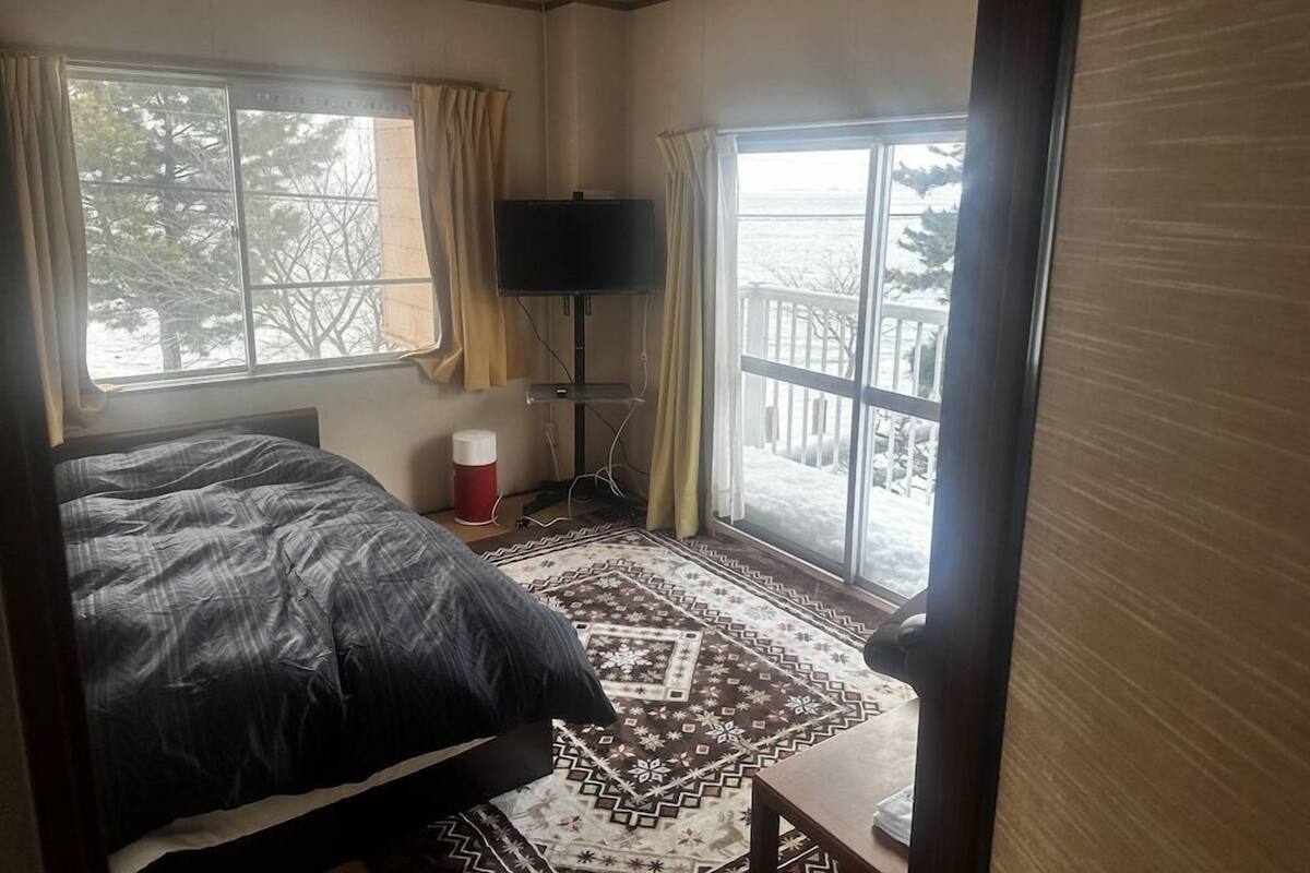 A bedroom with a bed, a TV, a window with curtains, and a view of the outdoors.