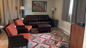 Ein Wohnzimmer mit einem braunen Ledersofa, roten und schwarzen Korbstühlen, einem gemusterten Teppich und einem Gemälde an der Wand.