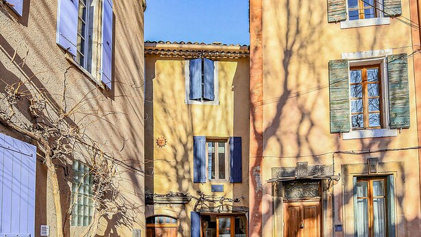 A narrow street with buildings featuring traditional architecture, including shuttered windows and a central door.
