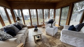 Una habitación acogedora con techo de madera, ventanas amplias, un sofá blanco, un sillón blanco y una pequeña mesa con un adorno.