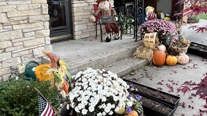 Una casa con fachada de piedra, un porche con un espantapájaros decorativo, y una variedad de macetas con plantas y calabazas.