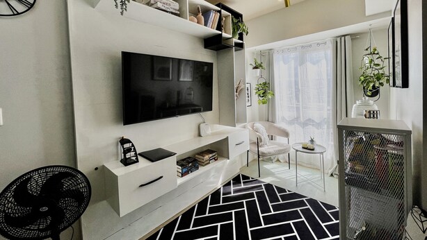 Una sala moderna con un televisor de pantalla plana, estanterías blancas, un tapete de diseño blanco y negro y una pequeña mesita con una planta.