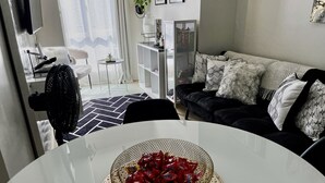 Un salón moderno con una mesa de comedor en primer plano, un sofá negro con cojines estampados y un dispensador de agua sobre un mueble blanco.