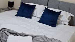 Un lit bien fait, avec une literie blanche et grise, deux oreillers bleus et une œuvre d’art abstraite encadrée accrochée au mur.