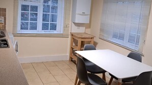 Une cuisine compacte avec un plan de travail blanc, une table en bois et des chaises. Il y a une fenêtre avec des stores et un meuble blanc au-dessus du plan de travail.