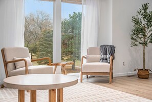 Een moderne woonkamer met twee beige fauteuils, een ronde houten tafel en uitzicht op bomen door grote ramen.