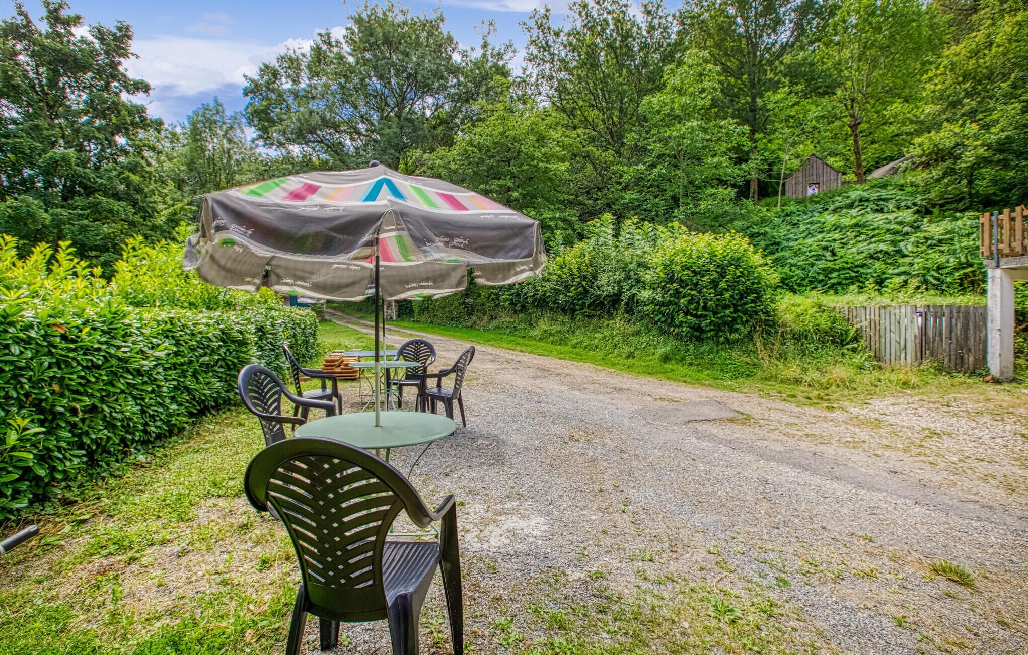 A gravel path leading to a covered outdoor seating area with a table and chairs, surrounded by lush greenery and a wooden structure in the background.