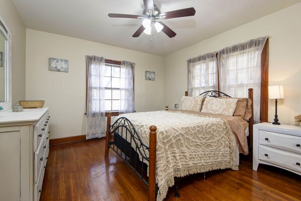 A bedroom with a bed, a nightstand, a dresser, a ceiling fan, and two windows with curtains.