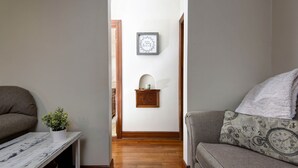 Un pasillo con piso de madera, una mesa blanca con una planta, un sofá gris con un cojín estampado y una pared decorada con cuadros.