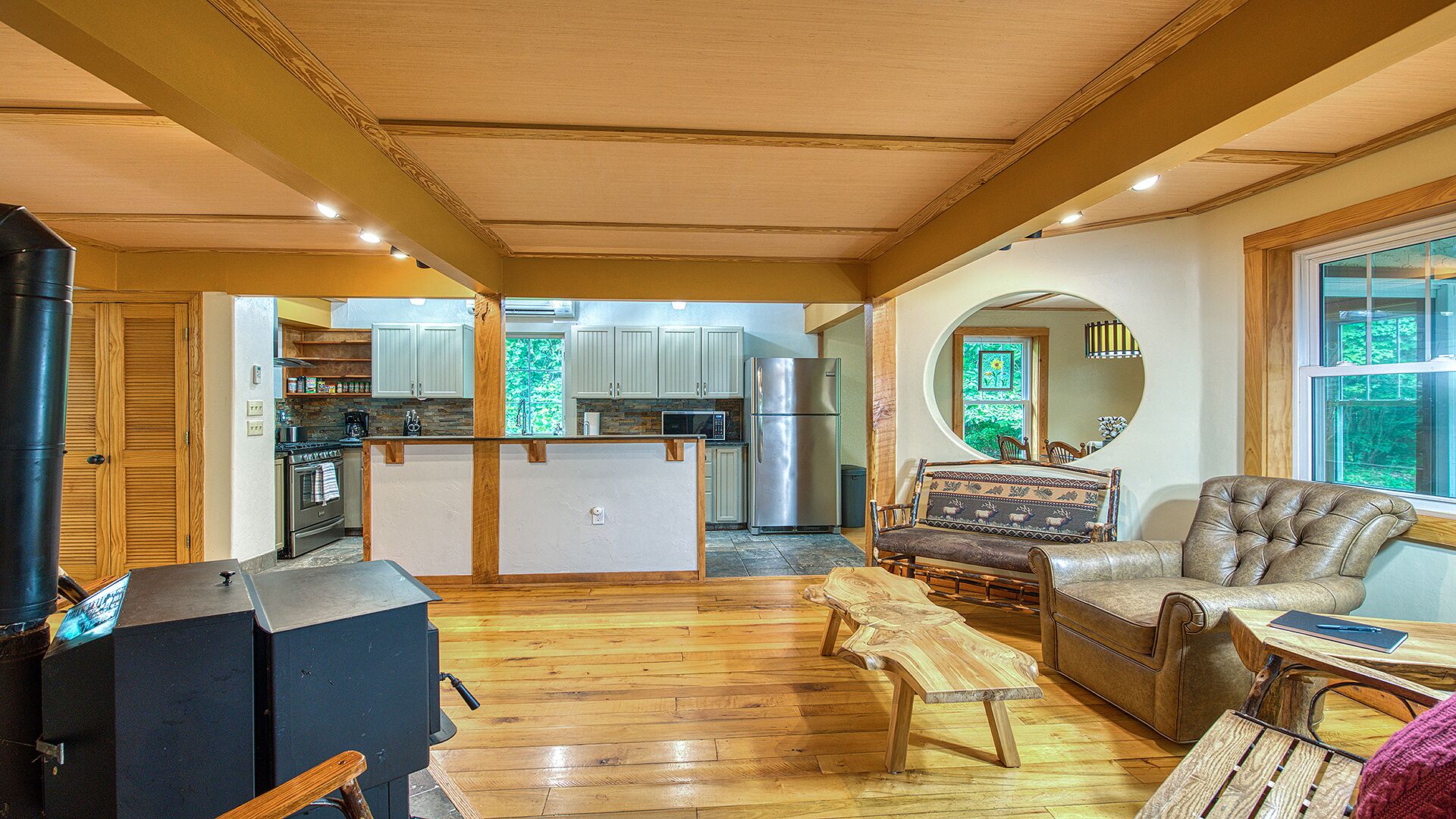 A spacious living area with a wooden floor, a kitchen with white cabinets, a large mirror, and a leather sofa.
