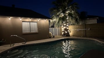 A swimming pool with a palm tree and string lights, surrounded by a brick wall and a window.
