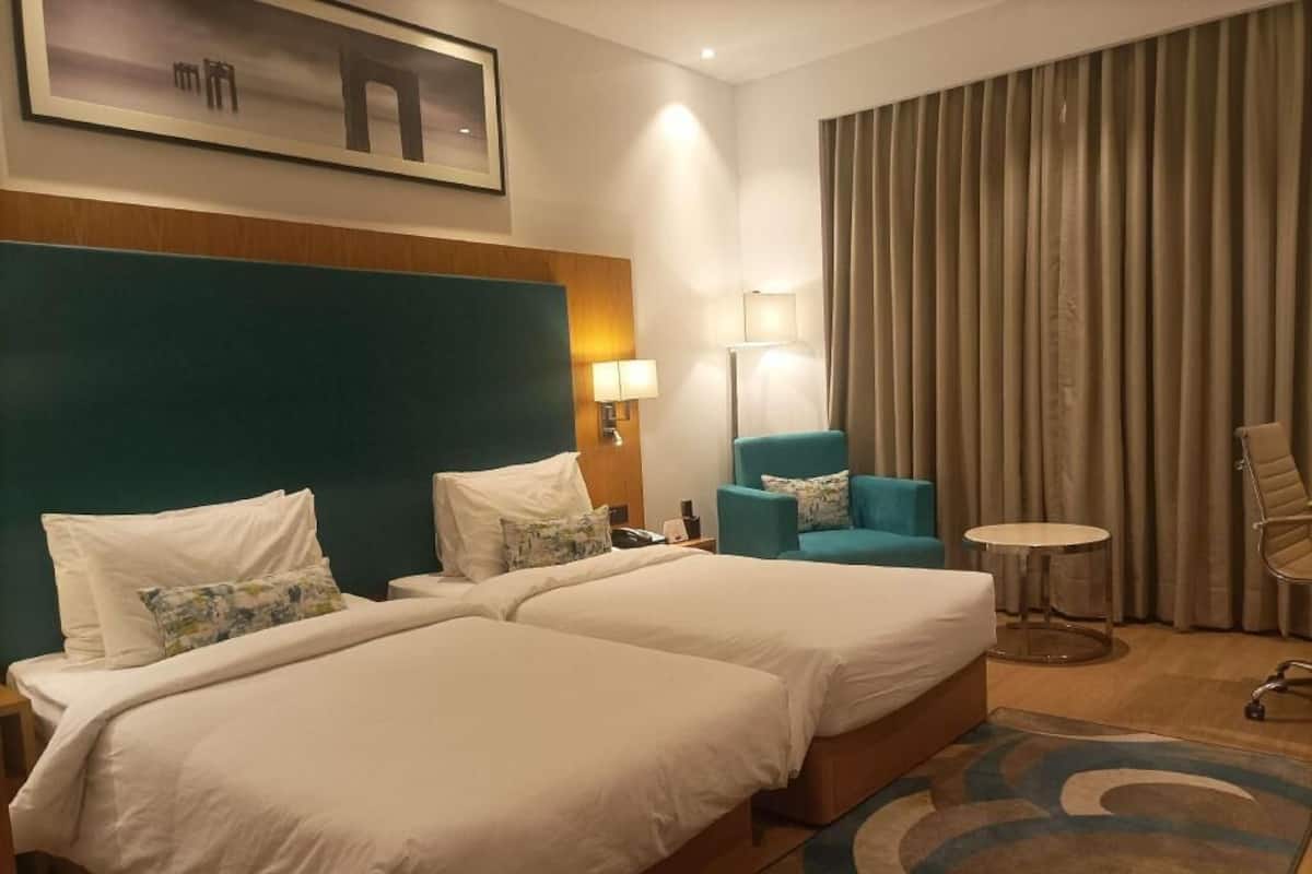 Quarto de hotel com duas camas, uma mesa, uma cadeira, uma luminária e uma janela grande com cortinas.