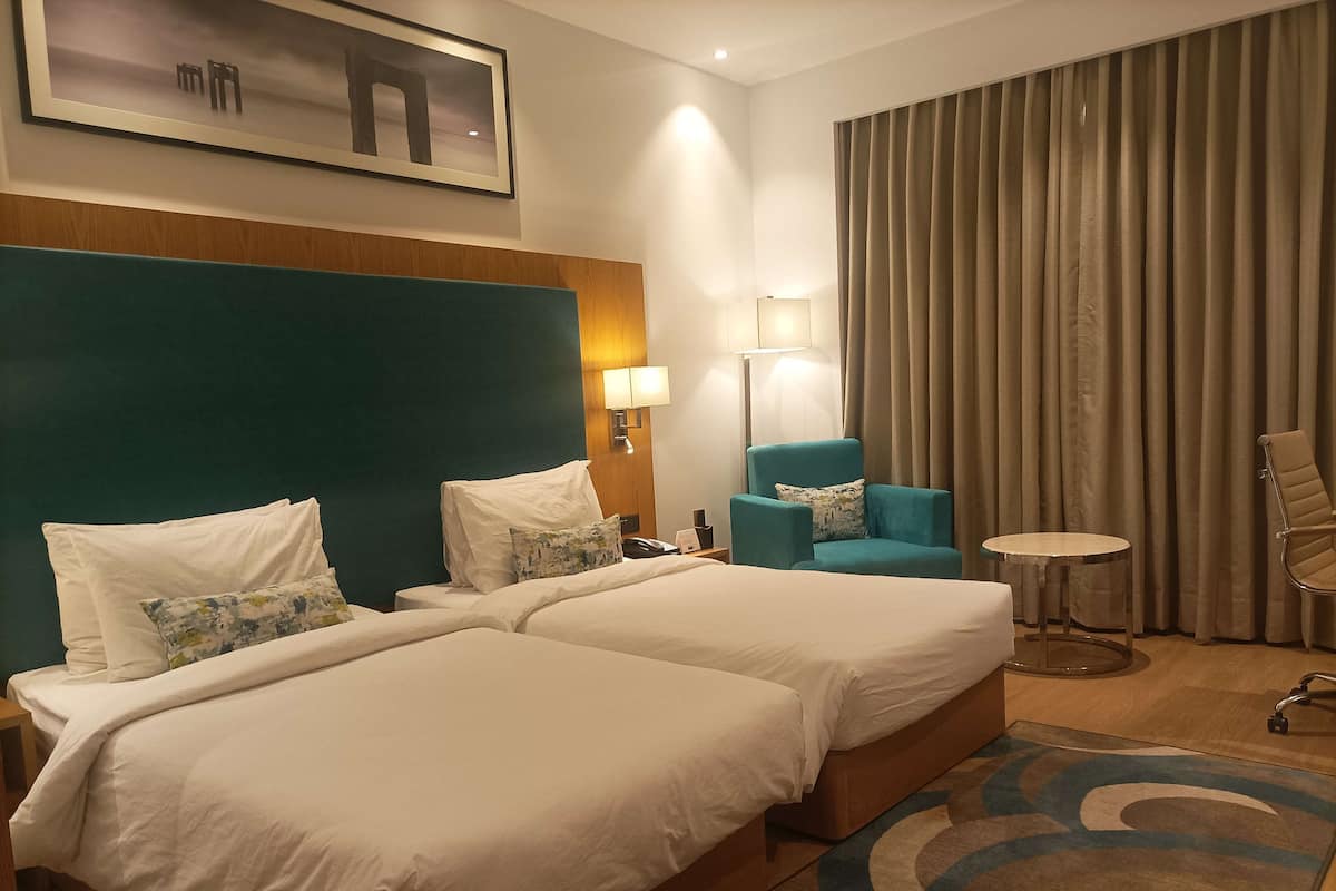 Quarto de hotel com duas camas, uma parede de destaque verde-água, uma mesa redonda pequena, uma cadeira e uma janela grande com cortinas.