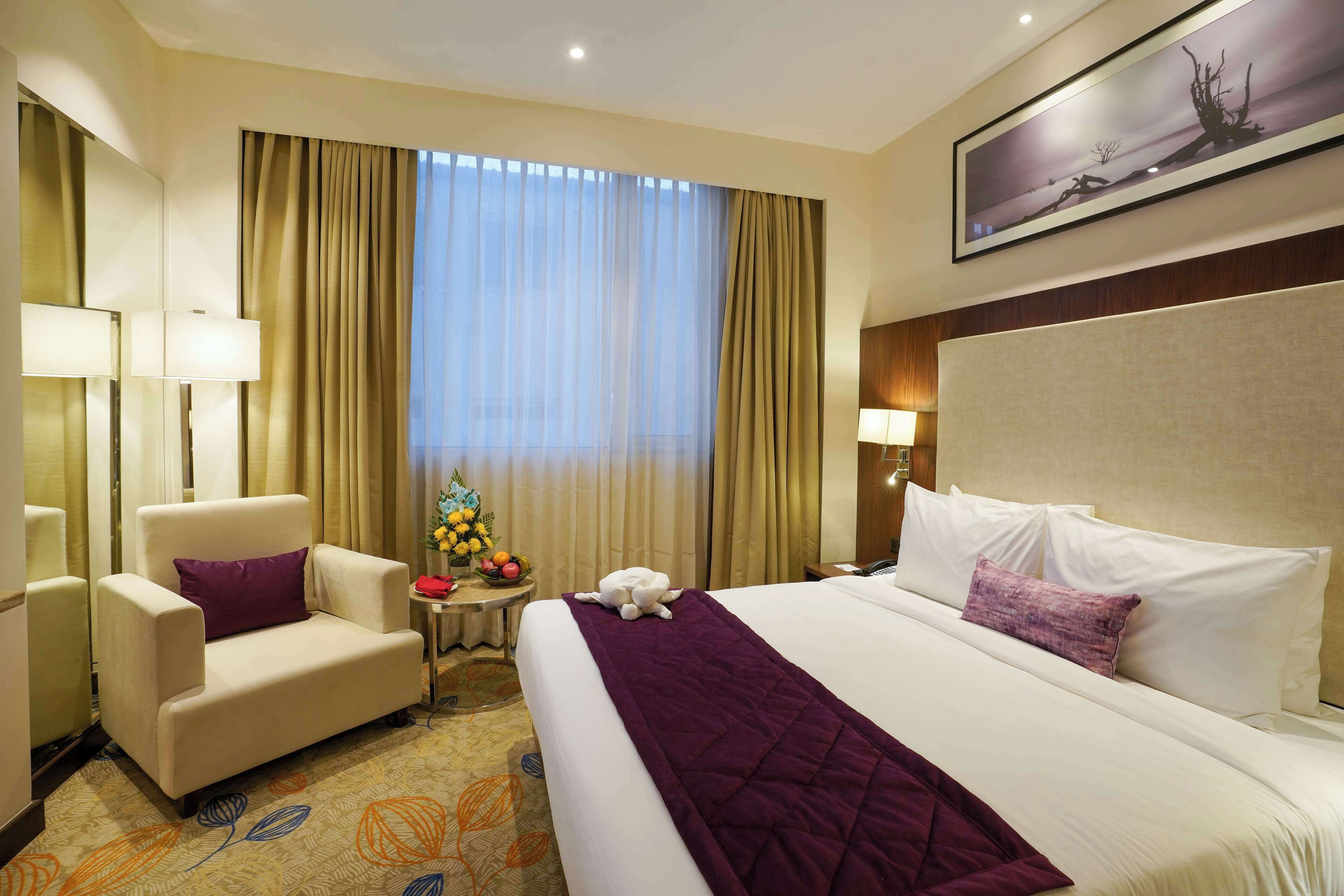 A hotel room with a large bed, a chair, a small table with a fruit bowl, and a painting on the wall.