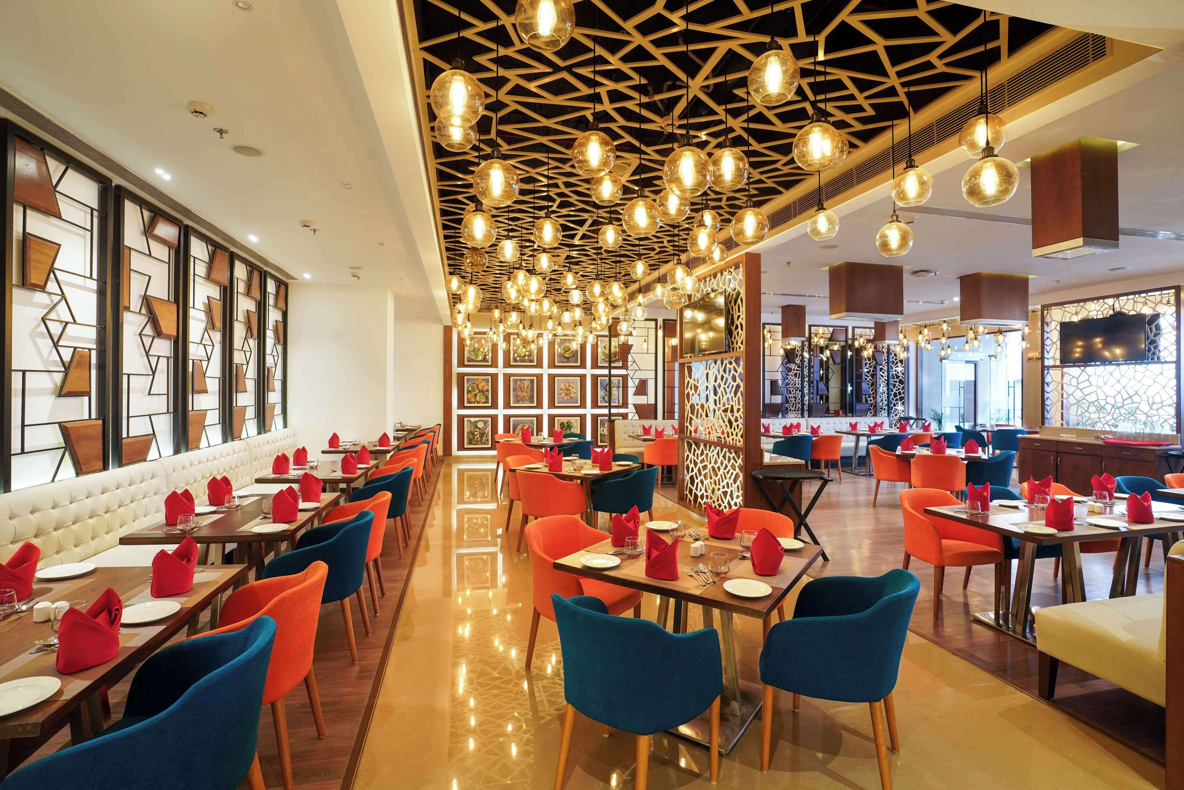 Un restaurante con una decoración moderna, que presenta una amplia zona de comedor con mesas y sillas, un muro con diseño y un techo con luces colgantes.