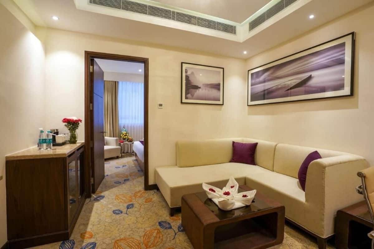 Quarto de hotel com sofá, mesa de centro, escrivaninha com um vaso de flores e duas fotos emolduradas na parede.
