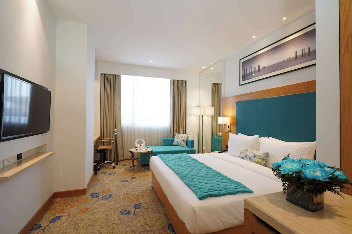 Quarto de hotel com uma cama grande, televisão, escrivaninha com abajur, cadeira e uma mesinha.