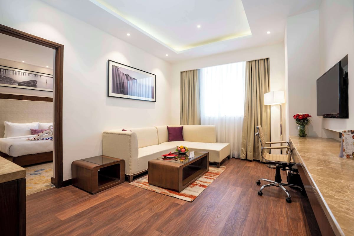 Quarto de hotel com cama, sofá, mesa de centro, televisão e escrivaninha com abajur.