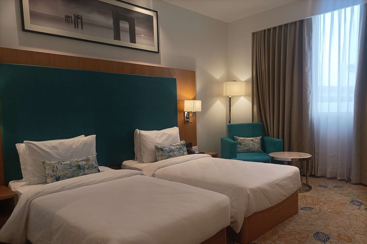 Quarto de hotel com duas camas, uma poltrona verde-água, uma mesinha redonda e uma luminária.